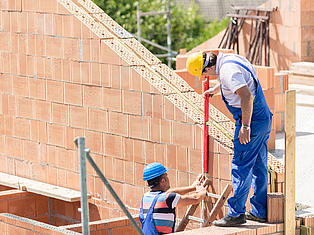 Zwei Bauarbeiter mit Schutzhelmen arbeiten auf einem Gerüst an einer im Bau befindlichen Ziegelwand.