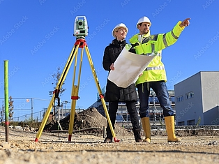 Zwei Bauarbeiter auf einer Baustelle prüfen mit einem Vermessungsgerät das Gelände; einer zeigt in die Ferne.