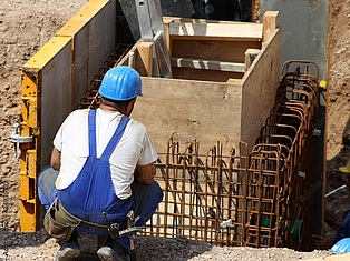 Bauarbeiter mit blauem Schutzhelm kniet neben einer Bewehrung aus Stahlstäben auf einer Baustelle.
