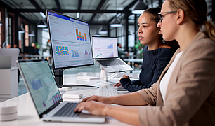 Zwei Frauen arbeiten an Laptops und betrachten Datenanalysen mit Diagrammen auf mehreren Bildschirmen.